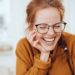 Pretty red-haired girl with pigtail, wearing glasses and orange sweatshirt, laughing