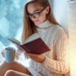 woman in glasses in knitted winter white warm sweater drinks a cup of hot cocoa during reading favorite book on a window sill by the window at home in winter