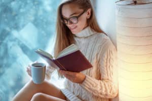 woman in glasses in knitted winter white warm sweater drinks a cup of hot cocoa during reading favorite book on a window sill by the window at home in winter