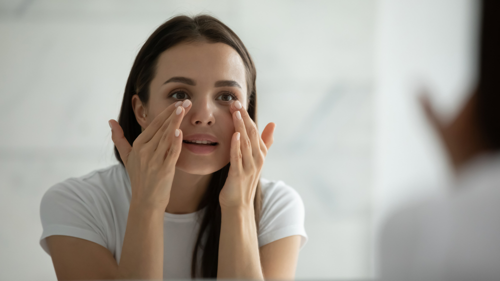 Beautiful young woman touching her under eye in the mirror after getting dermal fillers for eye hollows.